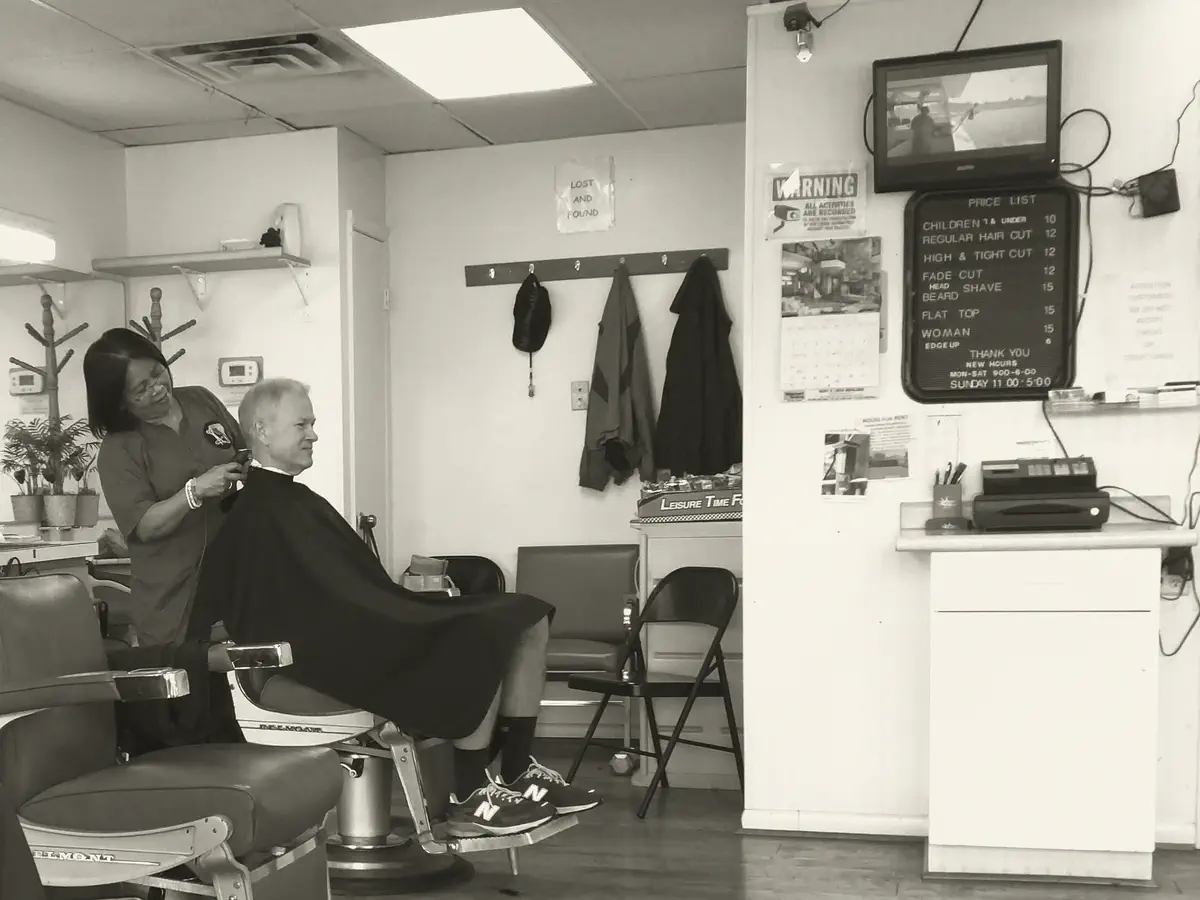 Inside the shop: a barber at work at a Belmont chair, coat hooks along the wall and the letter-board price list at the back